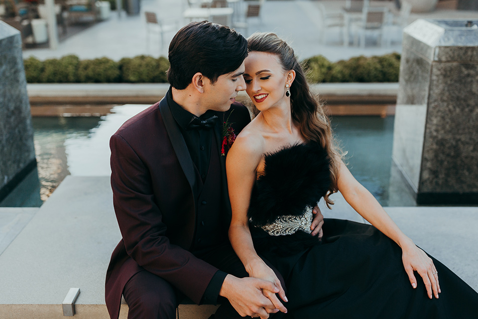 dia-de-los-muertos-shoot-bride-and-groom-bride-without-floral-headpiece-bride-and-grom-out-of-focus-a-little-smilingbride-in-a-black-dress-with-a-floral-headpeice-andbig-floral-bouquet-groom-in-burgundy-jacket-with-black-trim-black-shirt-and-a-faux-leather-bow-tie