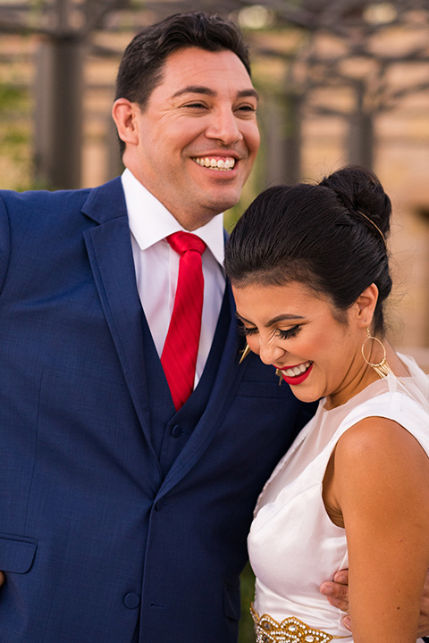 wonder-woman-meets-superman-laughing-bride-in-a-white-silk-dress-with-her-wonder-woman-head-piece-holding-onto-groom-in-a-navy-suit-and-red-long-tie-with-red-rose-florals