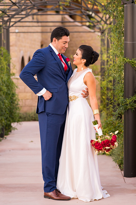 wonder-woman-meets-superman-looking-at-each-other-bride-in-a-white-silk-dress-with-her-wonder-woman-head-piece-holding-onto-groom-in-a-navy-suit-and-red-long-tie-with-red-rose-florals