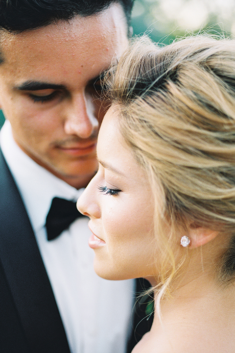 Inn-at-rancho-santa-fe-shoot-bride-and-groom-close-up-with-bride-in-foreground-with-back-towards-camera-looking-to-the-side-groom-in-background-in-focus-looking-at-her-Inn-at-rancho-santa-fe-shoot-bride-and-groom-looking-at-each-other-bride-in-a-lace-gown-with-an-illusion-detailing-with-a-nude-underlay-with-her-hair-in-back-in-a-loose-bun-groom-in-a-black-tuxedo-with-black-blow-tie-and-a-white-and-black-dolka-dot-pocket-square