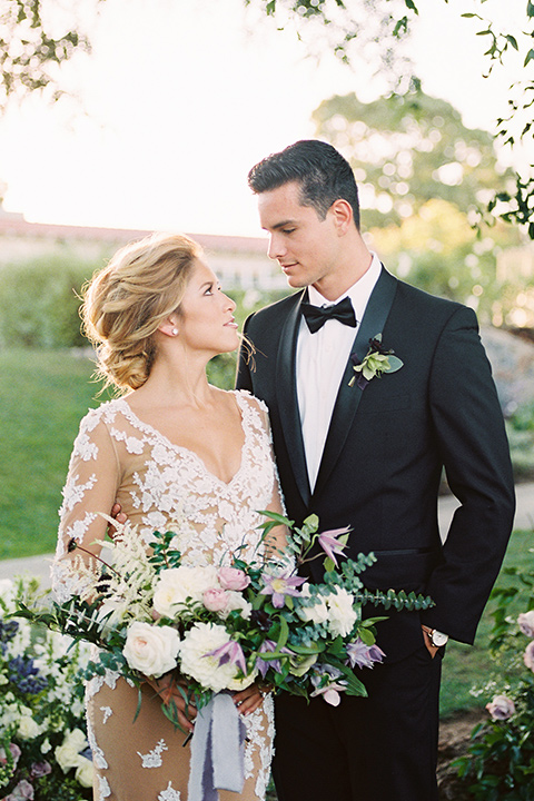 Inn-at-rancho-santa-fe-shoot-bride-and-groom-looking-at-each-other-holding-flowers-by-the-tree-close-up-bride-in-a-lace-gown-with-an-illusion-detailing-with-a-nude-underlay-with-her-hair-in-back-in-a-loose-bun-groom-in-a-black-tuxedo-with-black-blow-tie-and-a-white-and-black-olka-dot-pocket-square