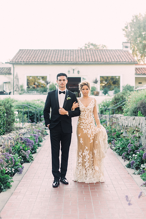 Inn-at-rancho-santa-fe-shoot-bride-and-groom-standing-in-garden-walk-way-holding-hands-bride-in-a-lace-gown-with-an-illusion-detailing-with-a-nude-underlay-with-her-hair-in-back-in-a-loose-bun-groom-in-a-black-tuxedo-with-black-blow-tie-and-a-white-and-black-polka-dot-pocket-square