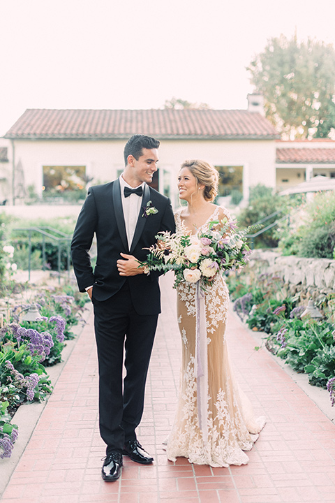 Inn-at-rancho-santa-fe-shoot-bride-and-groom-walking-bride-in-a-lace-gown-with-an-illusion-detailing-with-a-nude-underlay-with-her-hair-in-back-in-a-loose-bun-groom-in-a-black-tuxedo-with-black-blow-tie-and-a-white-and-black-olka-dot-pocket-square