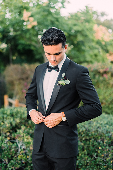 Inn-at-rancho-santa-fe-shoot-groom-buttoning-jacket-groom-along-standing-looking-straight-on-camera-groom-in-a-black-tuxedo-with-black-blow-tie-and-a-white-and-black-polka-dot-pocket-square