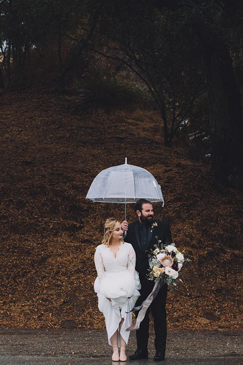 amanda-and-codys-wedding-at-the-1909-bride-and-groom-crossing-street-with-umbrella-bride-in-an-a-line-dress-with-a-tulle-skirt-and-lace-long-sleeve-bodice-groom-in-all-black-shawl-lapel-tuxedo