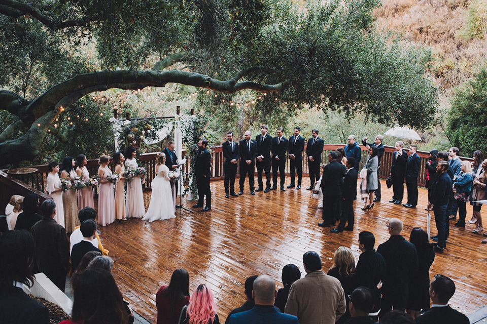 amanda-and-codys-wedding-at-the-1909-ceremony-space-bride-in-an-a-line-dress-with-a-tulle-skirt-and-lace-long-sleeve-bodice-groom-in-all-black-shawl-lapel-tuxedo-rainy-day-all-guests-in-a-half-moon-shape-around-them