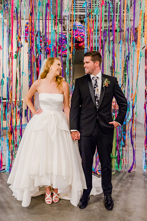 Childrens-museum-shoot-bride-and-groom-colorful-ribbon-backdrop-bride-in-a-white-modern-strapless-ball-gown-and-groom-in-a-black-tuxedo-with-a-floral-tie