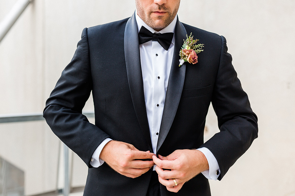 Childrens-museum-shoot-groom-buttoning-jacket-groom-in-a-black-tuxedo-with-a-floral-tie