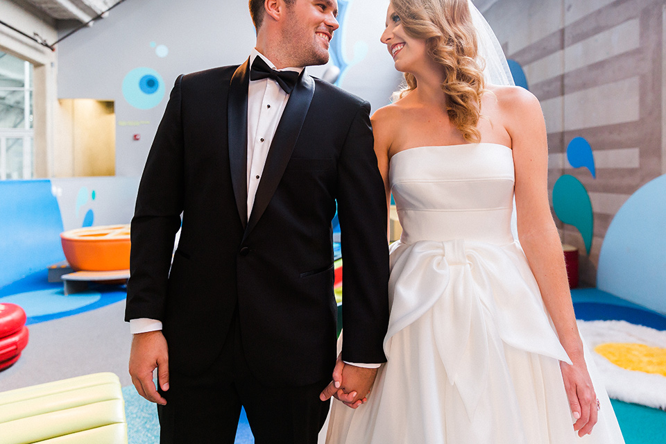 childerns-museum-bride-and-groom-close-up-looking-at-them-bride-and-groom-colorful-ribbon-backdrop-bride-in-a-white-modern-strapless-ball-gown-and-groom-in-a-black-tuxedo-with-a-floral-tie