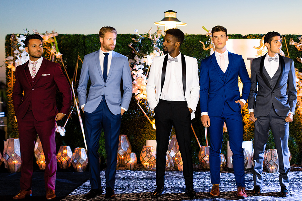 evolution-of-style-party-burgundy-model-on-the-end-guys-showing-off-latest-styles-starting-from-the-left-model-in-burgundy-suit-with-white-and-burgundy-floral-tie-next-model-in-dark-blue-ants-with-a-light-blue-suit-jacket-and-navy-tie-next-model-in-a-white-juxedo-jacket-with-black-trim-and-black-pants-next-model-in-cobalt-suit-with-blush-tie-and-brown-shoes-and-last-model-in-grey-tuxedo-with-black-trim at-the-fifth-restaurant