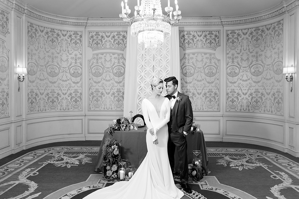 Exquisite-weddings-magazine-black-and-white-photo-of-bride-and-groom-at-sweetheart-table-bride-in-a-white-long-sleeved-dress-with-a-plunging-neckline-and-hair-in-a-bun-groom-in-a-grey-michael-kors-tuxedo-with-a-black-bowtie