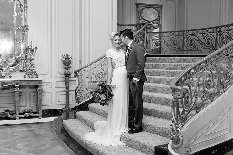 Exquisite-weddings-magazine-black-and-white-photo-on-staircase-bride-in-a-white-long-sleeved-dress-with-a-plunging-neckline-and-hair-in-a-bun-groom-in-a-grey-michael-kors-tuxedo-with-a-black-bowtie