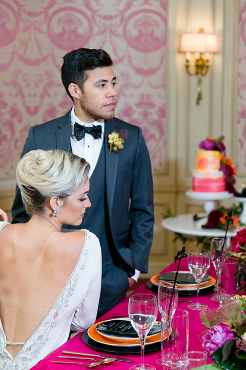 Exquisite-weddings-magazine-bride-and-groom-at-the-desserts-table-looking-away-bride-in-a-white-long-sleeved-dress-with-a-plunging-neckline-and-hair-in-a-bun-groom-in-a-grey-michael-kors-tuxedo-with-a-black-bowtie