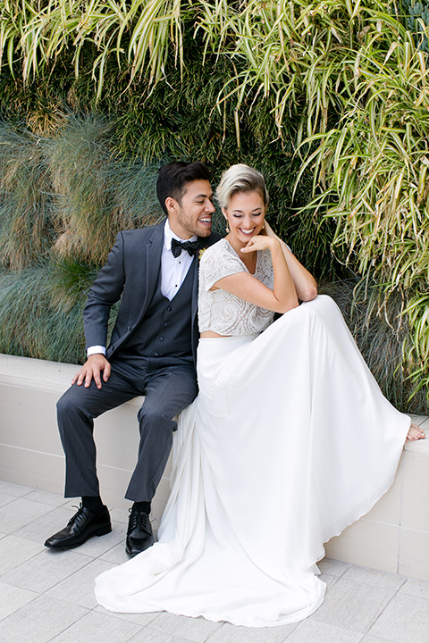 Exquisite-weddings-magazine-bride-and-groom-sitting-outside-laughing-bride-in-a-white-long-sleeved-dress-with-a-plunging-neckline-and-hair-in-a-bun-groom-in-a-grey-michael-kors-tuxedo-with-a-black-bowtie