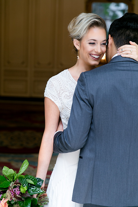 Exquisite-weddings-magazine-bride-holding-onto-groom-laughing-bride-in-a-white-long-sleeved-dress-with-a-plunging-neckline-and-hair-in-a-bun-groom-in-a-grey-michael-kors-tuxedo-with-a-black-bowtie