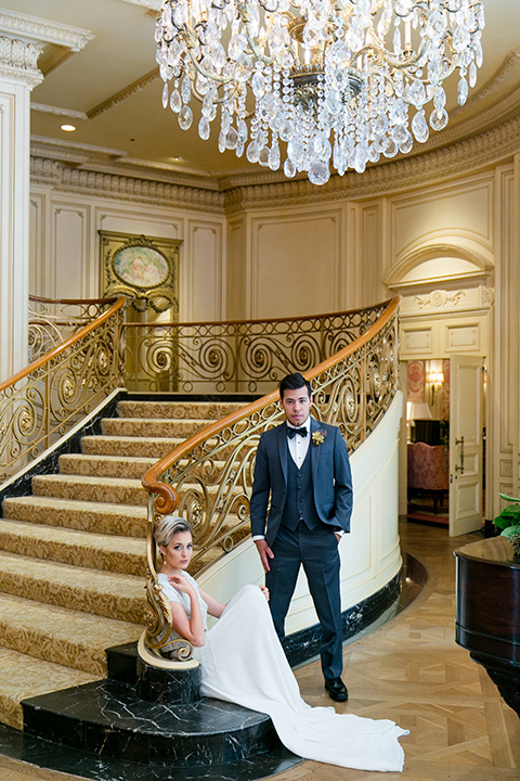 Exquisite-weddings-magazine-bride-sitting-on-stairs-groom-standing-next-to-her-bride-in-a-white-long-sleeved-dress-with-a-plunging-neckline-and-hair-in-a-bun-groom-in-a-grey-michael-kors-tuxedo-with-a-black-bowtie