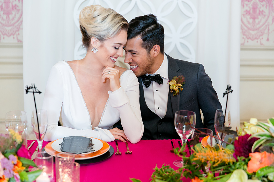 Exquisite-weddings-magazine-brode-and-groom-laughing-at-sweetheart-table-bride-in-a-white-long-sleeved-dress-with-a-plunging-neckline-and-hair-in-a-bun-groom-in-a-grey-michael-kors-tuxedo-with-a-black-bowtie