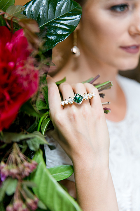 Exquisite-weddings-magazine-close-up-on-emerald-ring-bride-in-a-white-long-sleeved-dress-with-a-plunging-neckline-and-hair-in-a-bun-with-an-emerald-ring-and-a-bright-floral-arrangement