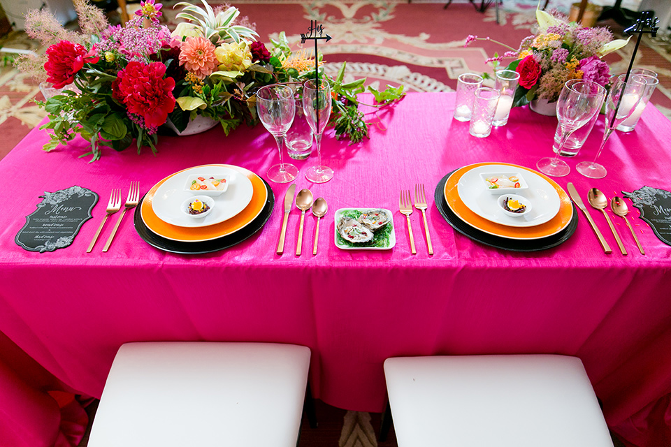 Exquisite-weddings-magazine-close-up-on-table-hot-pink-linen-with-white-plates-and-gold-cutlery-with-bright-orange-and-green-floral-arrangements