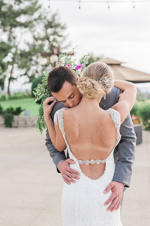 Temecula outdoor wedding at callaway winery bride form fitting lace gown with crystal beading and lace detail with open back design and beaded straps with groom grey notch lapel suit with matching vest and white dress shirt with long black skinny tie and pink floral boutonniere hugging