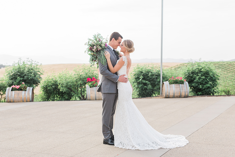 Temecula outdoor wedding at callaway winery bride form fitting lace gown with crystal beading and lace detail with open back design and beaded straps with groom grey notch lapel suit with matching vest and white dress shirt with long black skinny tie and pink floral boutonniere hugging and dancing bride holding pink and green floral bridal bouquet