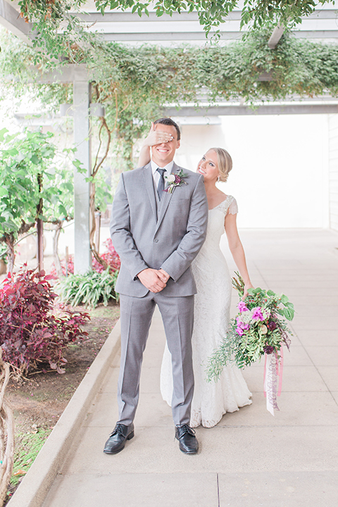 Temecula outdoor wedding at callaway winery bride form fitting lace gown with crystal beading and lace detail with open back design and beaded straps with groom grey notch lapel suit with matching vest and white dress shirt with long black skinny tie and pink floral boutonniere first look bride holding pink and green floral bridal bouquet