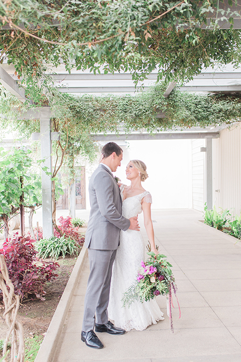 Temecula outdoor wedding at callaway winery bride form fitting lace gown with crystal beading and lace detail with open back design and beaded straps with groom grey notch lapel suit with matching vest and white dress shirt with long black skinny tie and pink floral boutonniere first look hugging and bride holding pink and green floral bridal bouquet