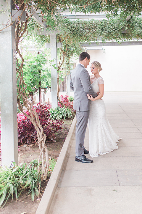 Temecula outdoor wedding at callaway winery bride form fitting lace gown with crystal beading and lace detail with open back design and beaded straps with groom grey notch lapel suit with matching vest and white dress shirt with long black skinny tie and pink floral boutonniere hugging first look bride holding pink and green floral bridal bouquet