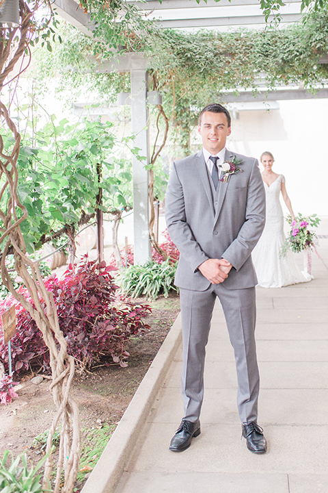 Temecula outdoor wedding at callaway winery bride form fitting lace gown with crystal beading and lace detail with open back design and beaded straps with groom grey notch lapel suit with matching vest and white dress shirt with long black skinny tie and pink floral boutonniere first look bride holding pink and green floral bridal bouquet