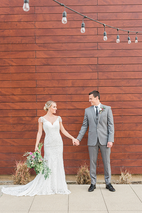 Temecula outdoor wedding at callaway winery bride form fitting lace gown with crystal beading and lace detail with open back design and beaded straps with groom grey notch lapel suit with matching vest and white dress shirt with long black skinny tie and pink floral boutonniere holding hands and bride holding pink and green floral bridal bouquet