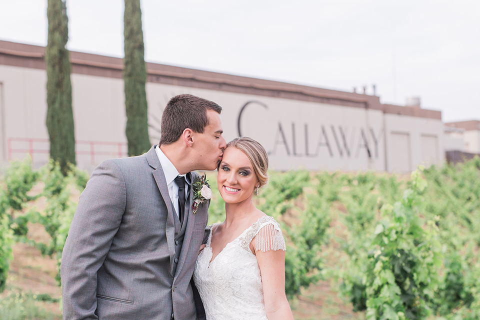 Temecula outdoor wedding at callaway winery bride form fitting lace gown with crystal beading and lace detail with open back design and beaded straps with groom grey notch lapel suit with matching vest and white dress shirt with long black skinny tie and pink floral boutonniere hugging and kissing bride holding pink and green floral bridal bouquet