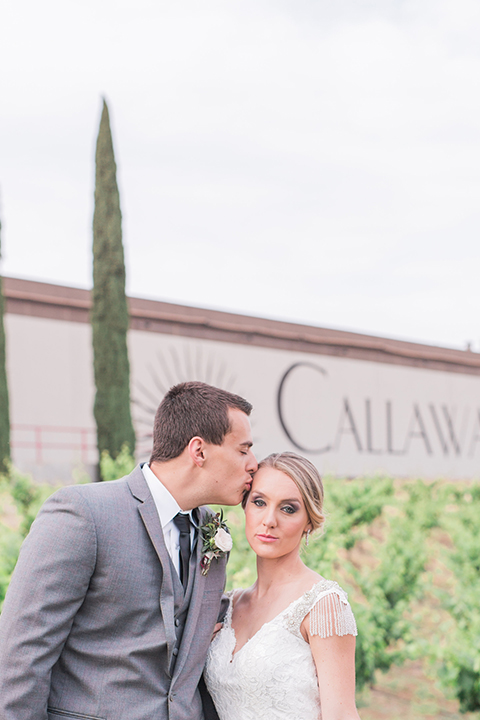 Temecula outdoor wedding at callaway winery bride form fitting lace gown with crystal beading and lace detail with open back design and beaded straps with groom grey notch lapel suit with matching vest and white dress shirt with long black skinny tie and pink floral boutonniere hugging and kissing bride holding pink and green floral bridal bouquet