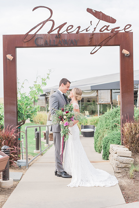 Temecula outdoor wedding at callaway winery bride form fitting lace gown with crystal beading and lace detail with open back design and beaded straps with groom grey notch lapel suit with matching vest and white dress shirt with long black skinny tie and pink floral boutonniere hugging and bride holding pink and green floral bridal bouquet