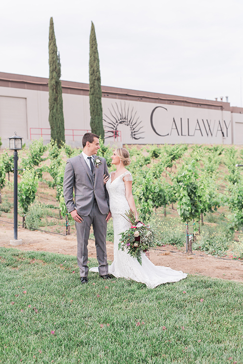 Temecula outdoor wedding at callaway winery bride form fitting lace gown with crystal beading and lace detail with open back design and beaded straps with groom grey notch lapel suit with matching vest and white dress shirt with long black skinny tie and pink floral boutonniere holding hands and bride holding pink and green floral bridal bouquet