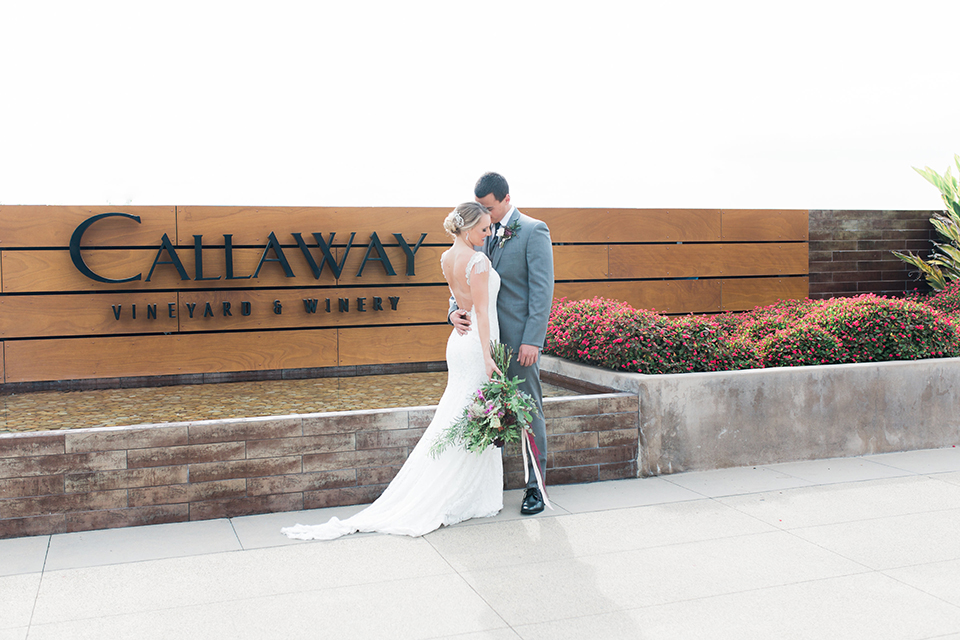 Temecula outdoor wedding at callaway winery bride form fitting lace gown with crystal beading and lace detail with open back design and beaded straps with groom grey notch lapel suit with matching vest and white dress shirt with long black skinny tie and pink floral boutonniere hugging and bride holding pink and green floral bridal bouquet