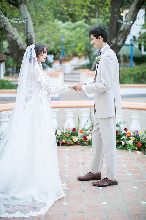 Rancho las lomas spanish inspired wedding shoot bride form fitting strapless lace gown with sweetheart neckline and long veil with lace trim and crystal belt with groom tan suit with matching vest and white dress shirt with dark grey bow tie holding hands