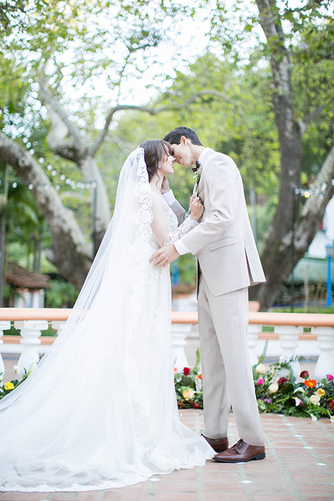 Rancho las lomas spanish inspired wedding shoot bride form fitting strapless lace gown with sweetheart neckline and long veil with lace trim and crystal belt with groom tan suit with matching vest and white dress shirt with dark grey bow tie kissing