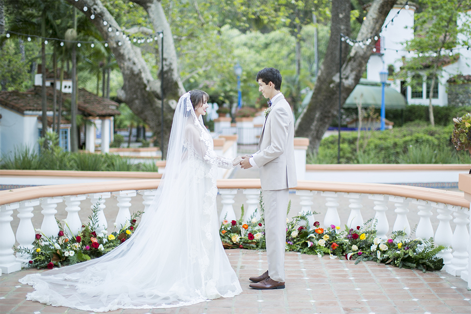 Rancho las lomas spanish inspired wedding shoot bride form fitting strapless lace gown with sweetheart neckline and long veil with lace trim and crystal belt with groom tan suit with matching vest and white dress shirt with dark grey bow tie holding hands