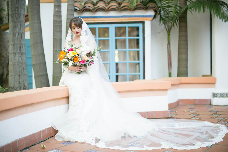 Rancho las lomas spanish inspired wedding shoot bride form fitting strapless lace gown with sweetheart neckline and long veil with lace trim and crystal belt holding floral bridal bouquet