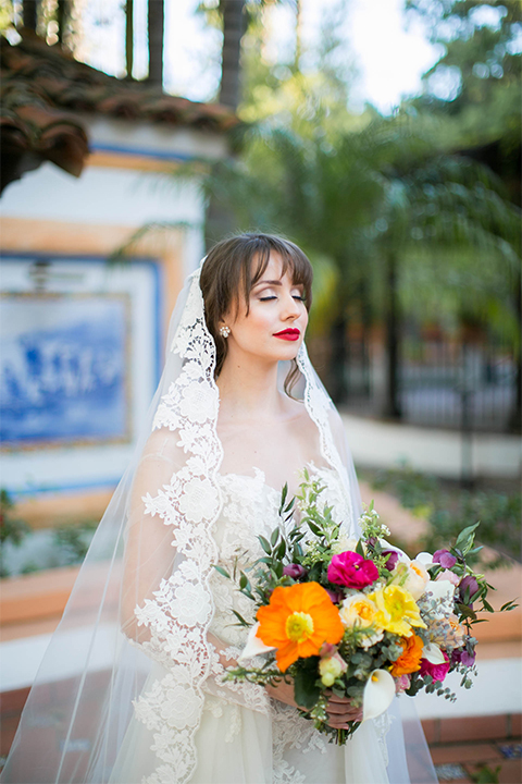 Rancho las lomas spanish inspired wedding shoot bride form fitting strapless lace gown with sweetheart neckline and long veil with lace trim and crystal belt holding bright floral bridal bouquet