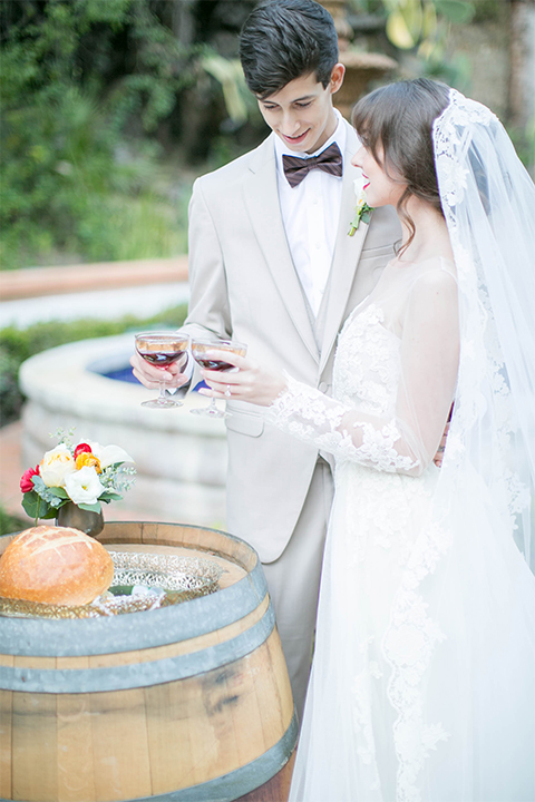 Rancho las lomas spanish inspired wedding shoot bride form fitting strapless lace gown with sweetheart neckline and long veil with lace trim and crystal belt with groom tan suit with matching vest and white dress shirt with dark grey bow tie holding glasses during ceremony