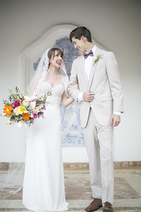 Rancho las lomas spanish inspired wedding shoot bride form fitting strapless lace gown with sweetheart neckline and long veil with lace trim and crystal belt with groom tan suit with matching vest and white dress shirt with dark grey bow tie holding grooms arm with bright colored floral bridal bouquet