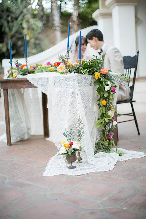 Rancho las lomas spanish inspired wedding shoot bride form fitting strapless lace gown with sweetheart neckline and long veil with lace trim and crystal belt with groom tan suit with matching vest and white dress shirt with dark grey bow tie sitting at table