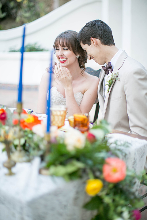 Rancho las lomas spanish inspired wedding shoot bride form fitting strapless lace gown with sweetheart neckline and long veil with lace trim and crystal belt with groom tan suit with matching vest and white dress shirt with dark grey bow tie sitting at table