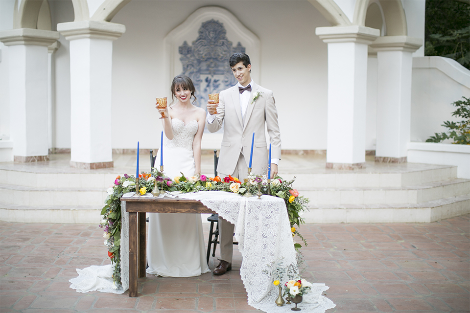 Rancho las lomas spanish inspired wedding shoot bride form fitting strapless lace gown with sweetheart neckline and long veil with lace trim and crystal belt with groom tan suit with matching vest and white dress shirt with dark grey bow tie standing by table