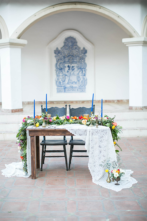 Rancho las lomas spanish inspired wedding shoot table set up with dark brown wood table and white lace table runner with green pink and orange flower centerpiece decor with tall blue candles and chairs