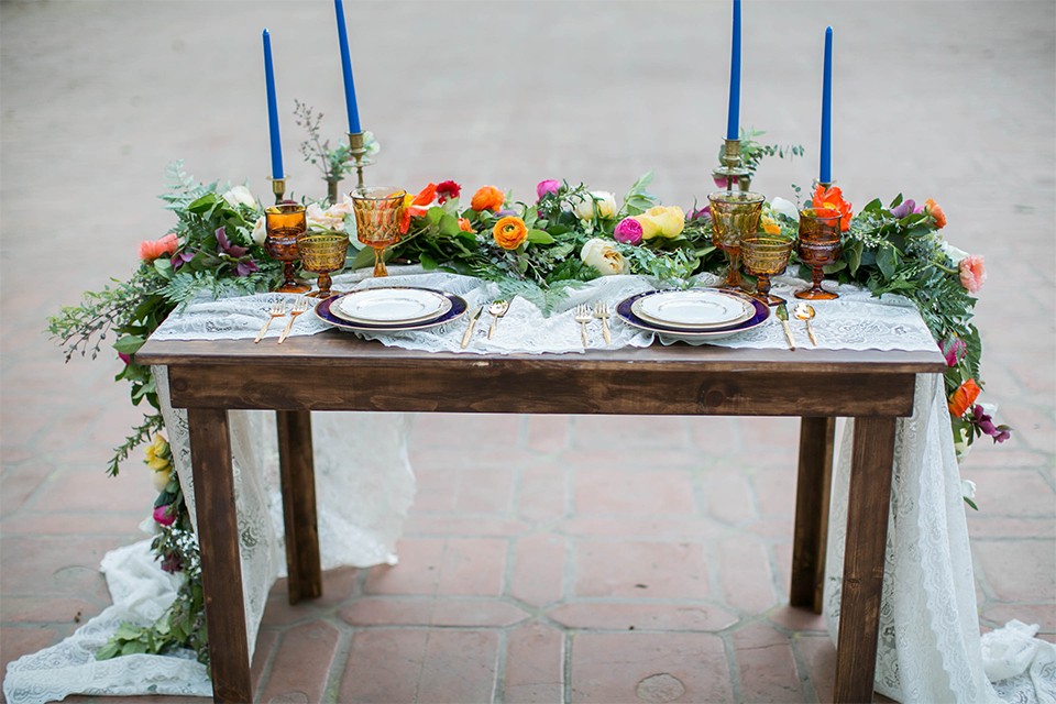 Rancho las lomas spanish inspired wedding shoot table set up with dark brown wood table and white lace table runner with green pink and orange flower centerpiece decor with tall blue candles and chairs