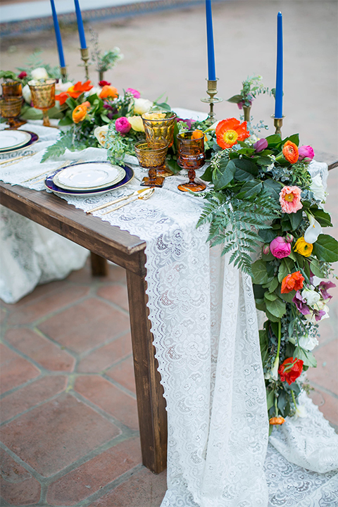 Rancho las lomas spanish inspired wedding shoot table set up with dark brown wood table and white lace table runner with green pink and orange flower centerpiece decor with tall blue candles and chairs