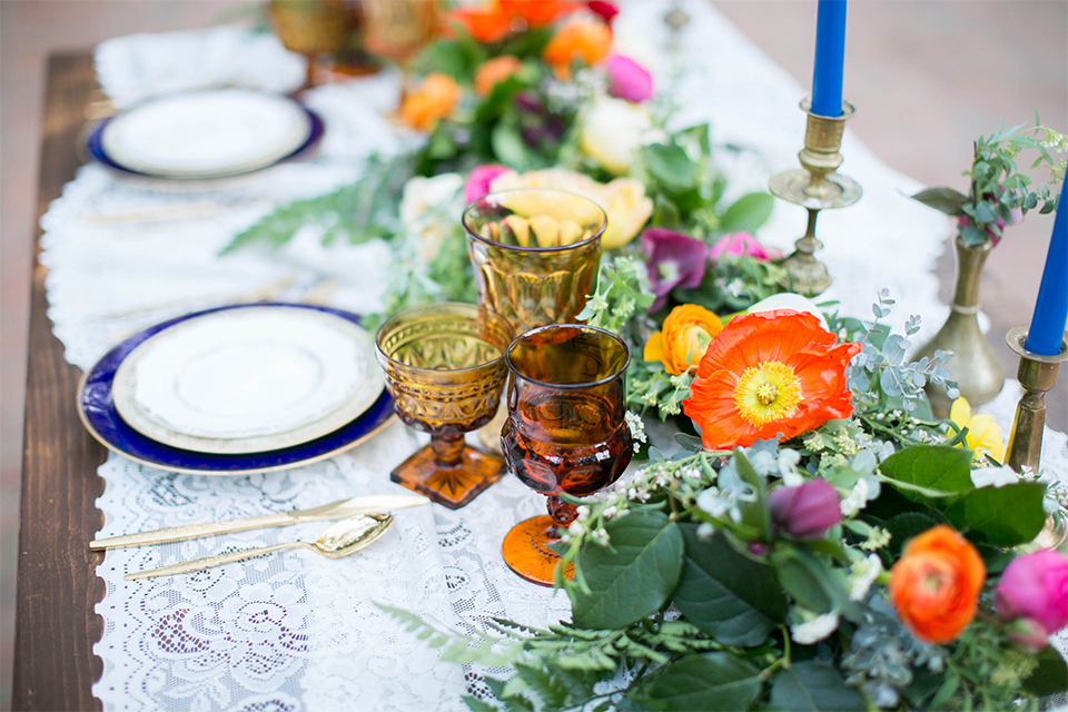 Rancho las lomas spanish inspired wedding shoot table set up with dark brown wood table and white lace table runner with green pink and orange flower centerpiece decor with tall blue candles and chairs