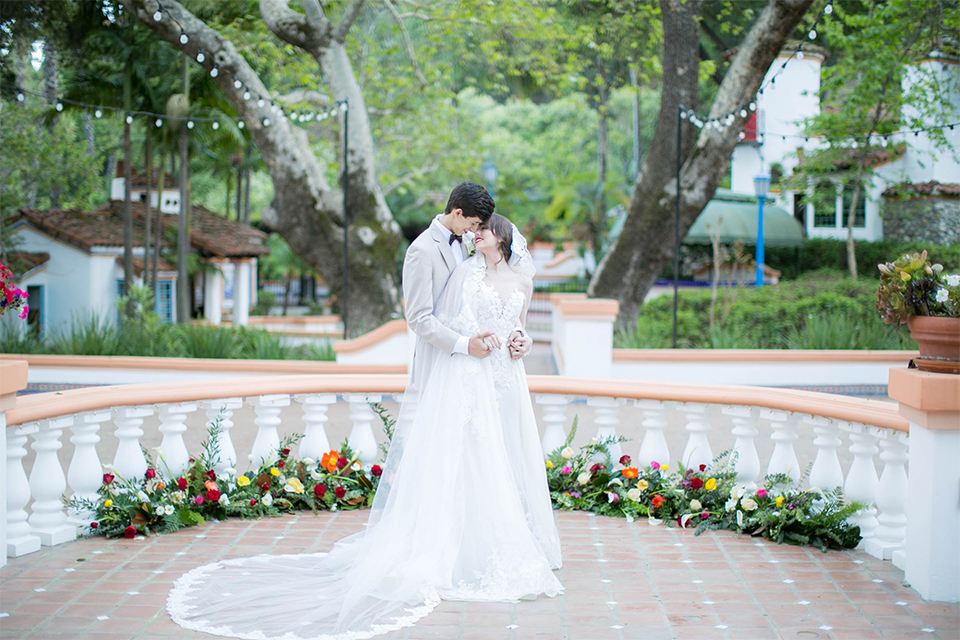 Rancho las lomas spanish inspired wedding shoot bride form fitting strapless lace gown with sweetheart neckline and long veil with lace trim and crystal belt with groom tan suit with matching vest and white dress shirt with dark grey bow tie hugging